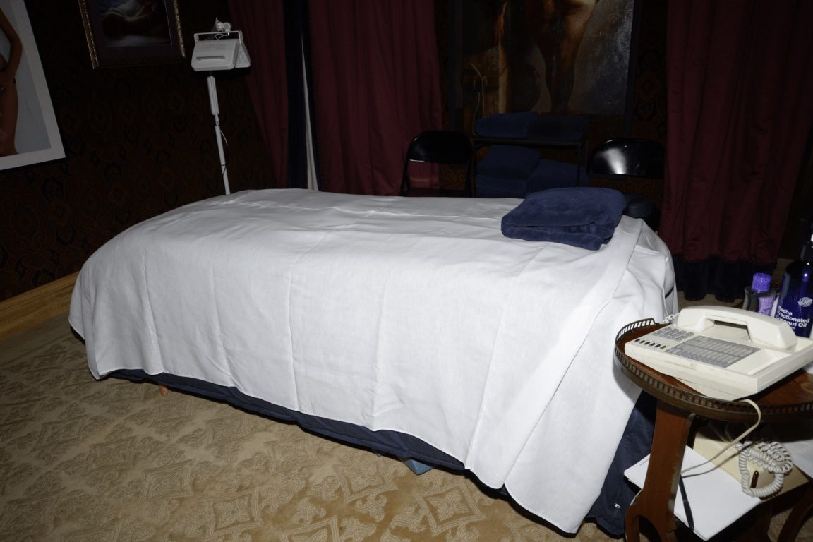 EFTA00000483 - Massage bed covered in white sheets with a blue towel on the headrest sits in a dimly lit room with red curtains and an old-fashioned telephone on a wooden side table